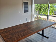 A wooden conference table with a dark, textured surface stands in a well-lit room with light gray walls and hardwood floors. Sunlight streams in through large windows with roller blinds partially down. A framed poster is on the wall.