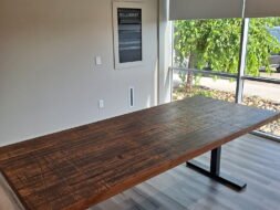 A wooden conference table with a dark, textured surface stands in a well-lit room with light gray walls and hardwood floors. Sunlight streams in through large windows with roller blinds partially down. A framed poster is on the wall.