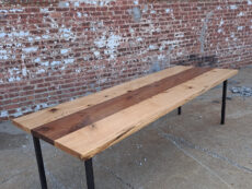 A long wooden table with metal legs stands on a concrete surface against a brick wall with several windows. The tabletop features a mix of light and dark wood tones, highlighting its natural grain.