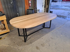 A wooden oval table with a light finish and black metal legs stands on a concrete floor in a workshop setting. Shelves and assorted industrial equipment are visible in the background.