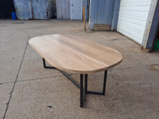 A wooden table with a smooth, oval-shaped top and metal legs is placed on a concrete surface outside a building. The background features industrial elements like corrugated metal walls and a partially open garage door.