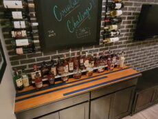 A shelf filled with various bottles of liquor sits against a brick wall. Above it, a chalkboard displays 2022 with colorful writing, and several wine bottles are mounted on racks on the side. A TV screen is partially visible on the right.