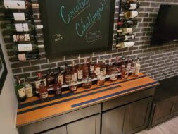 A shelf filled with various bottles of liquor sits against a brick wall. Above it, a chalkboard displays 2022 with colorful writing, and several wine bottles are mounted on racks on the side. A TV screen is partially visible on the right.