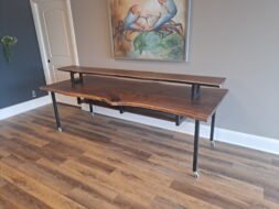 A wooden console table with a dark finish features a shelf supported by metal brackets. The table rests on wheels and stands on a wooden floor. A painting with birds is mounted on the wall above the table.