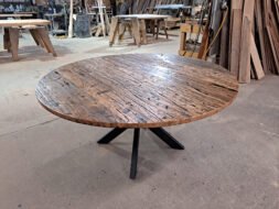A round wooden table with a rustic, reclaimed wood surface and a black, metal tripod base stands in a workshop setting. Surrounding the table are various woodworking tools and pieces of lumber, adding to the industrial atmosphere.
