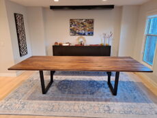 A dining room features a long wooden table with black metal legs on a blue patterned rug. In the background, there’s a sideboard with decorative items and a colorful landscape painting on a neutral wall. A window lets in natural light.