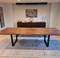 A dining room features a long wooden table with black metal legs on a blue patterned rug. In the background, there’s a sideboard with decorative items and a colorful landscape painting on a neutral wall. A window lets in natural light.