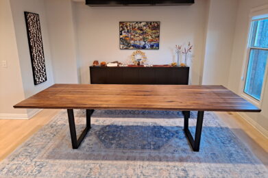 A dining room features a long wooden table with black metal legs on a blue patterned rug. In the background, there’s a sideboard with decorative items and a colorful landscape painting on a neutral wall. A window lets in natural light.