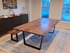 A modern dining room featuring a large wooden table with black metal legs and a matching bench. The room has light-colored walls, a large window, a blue patterned rug, and a sideboard topped with decor. A painting hangs above the sideboard.