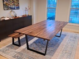 A modern dining room featuring a large wooden table with black metal legs and a matching bench. The room has light-colored walls, a large window, a blue patterned rug, and a sideboard topped with decor. A painting hangs above the sideboard.