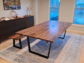 A modern dining room featuring a large wooden table with black metal legs and a matching bench. The room has light-colored walls, a large window, a blue patterned rug, and a sideboard topped with decor. A painting hangs above the sideboard.