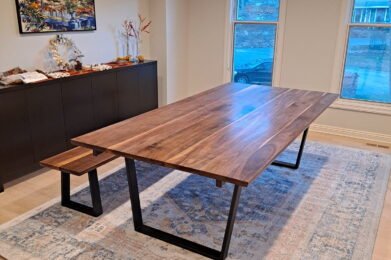 A modern dining room featuring a large wooden table with black metal legs and a matching bench. The room has light-colored walls, a large window, a blue patterned rug, and a sideboard topped with decor. A painting hangs above the sideboard.
