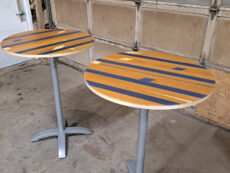 Two round tables with striped wooden tops and silver metal bases stand on a concrete floor. The background features a garage door with wooden panels.