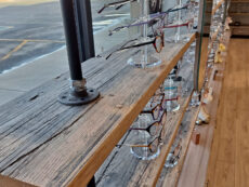 A wooden shelf with metal supports displays various eyeglasses on clear stands. The shelf is in front of a large window, revealing a parking lot and barren trees outside. The room has wooden flooring.