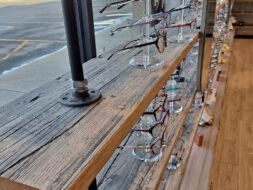 A wooden shelf with metal supports displays various eyeglasses on clear stands. The shelf is in front of a large window, revealing a parking lot and barren trees outside. The room has wooden flooring.
