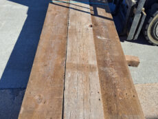 A long, weathered wooden plank is placed on a concrete surface. Shadows of a nearby forklift partially cover the plank, creating a pattern on its surface.