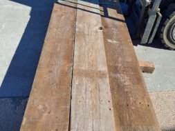 A long, weathered wooden plank is placed on a concrete surface. Shadows of a nearby forklift partially cover the plank, creating a pattern on its surface.
