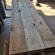 A long wooden table made of unfinished planks is positioned in a sunlit workshop. The surface has visible knots and grain patterns, and a strap partially secures the planks together, resting on a stack of additional wood.