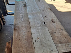 A long wooden table made of unfinished planks is positioned in a sunlit workshop. The surface has visible knots and grain patterns, and a strap partially secures the planks together, resting on a stack of additional wood.