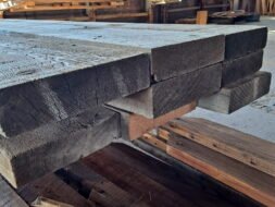 Stacks of large, weathered wooden beams are neatly piled in a warehouse. Sunlight streams in from windows, highlighting the rough and textured surfaces. The beams are arranged on a pallet, emphasizing the rustic, industrial setting.