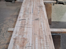 A long wooden table in a workshop setting, with visible wood grain and sawdust scattered on the floor. There are several tools and a blue chair in the background. The table is supported by makeshift wooden legs.