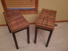 Two wooden side tables with dark metal legs are placed on a beige carpet. They are positioned at an angle, with a window with wooden blinds in the background, set against a light-colored wall.