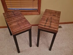 Two wooden side tables with dark metal legs are placed on a beige carpet. They are positioned at an angle, with a window with wooden blinds in the background, set against a light-colored wall.