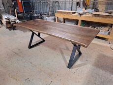 A rectangular wooden table with a natural finish and black metal legs in a workshop setting. Surrounding the table are various tools and materials, indicating an active woodworking environment.