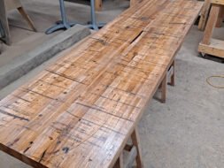 A large wooden table with a rustic finish is placed in a workshop. The table top shows natural grain patterns and signs of wear. Surrounding it are various woodworking tools and wooden planks. Sawdust is scattered on the floor.