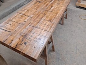 A large wooden table with a rustic finish is placed in a workshop. The table top shows natural grain patterns and signs of wear. Surrounding it are various woodworking tools and wooden planks. Sawdust is scattered on the floor.