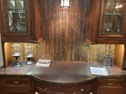A wooden cabinet with glass-fronted storage and a curved counter displays wine bottles on lower shelves. It features decorative lighting, wooden paneling, and a small book, blending rustic and elegant design elements.