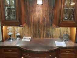 A wooden cabinet with glass-fronted storage and a curved counter displays wine bottles on lower shelves. It features decorative lighting, wooden paneling, and a small book, blending rustic and elegant design elements.
