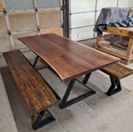 A rustic wooden table with a live edge design is flanked by two matching benches. The set has sturdy black metal legs and is situated in a garage workshop setting. A wooden workbench and a covered object are in the background.