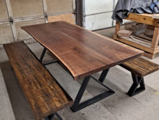 A rustic wooden table with a live edge design is flanked by two matching benches. The set has sturdy black metal legs and is situated in a garage workshop setting. A wooden workbench and a covered object are in the background.