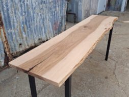 A long wooden table with a natural live edge sits on a concrete surface. The table has black metal legs and is set against a backdrop of weathered metal walls and a door. The wood grain is visible, showcasing its raw beauty.