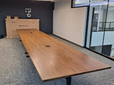 A modern conference room with a long wooden table, surrounded by gray carpet and walls. Circular ceiling lights illuminate the space. Large windows provide natural light. A flat-screen TV box is visible near the back wall.