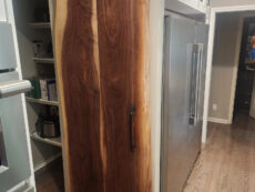 A kitchen features a sliding wooden barn door with a natural wood grain, separating the pantry area. Stainless steel appliances and white cabinets are visible, along with wooden flooring. The door is partially open, revealing pantry shelves.