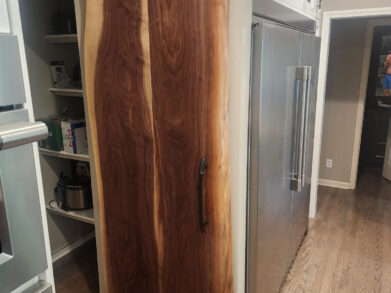 A kitchen features a sliding wooden barn door with a natural wood grain, separating the pantry area. Stainless steel appliances and white cabinets are visible, along with wooden flooring. The door is partially open, revealing pantry shelves.