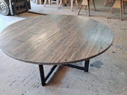 A large, round wooden table with a natural finish sits on a concrete floor in a spacious workshop. Various woodworking tools and materials are visible in the background, including a bandsaw and wooden planks.