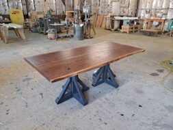 A large, rectangular wooden table with a smooth, polished surface sits on two sturdy blue trestle legs in a spacious workshop filled with various woodworking tools and equipment.