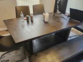 A dark wooden dining table with a matching bench and chairs, set with drinks, a water bottle, napkins, and a computer monitor, in a room with light-colored walls and windows with blinds.
