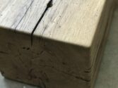 Close-up of a textured wooden beam with visible cracks and natural wood grain. The beam is situated on a light surface near a window, providing soft natural lighting.