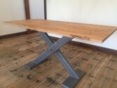 A wooden dining table with a smooth top and a modern metal X-shaped base, set on a rustic wooden floor in a room with white walls and wooden beams.