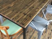 A wooden table with metal legs is surrounded by various chairs, including an upholstered wooden chair with a green cushion and several gray metal chairs. The floor has a rustic, scratched wooden appearance.