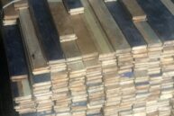 A stack of wooden planks is neatly piled up and strapped together with a white band. The planks vary in color, ranging from light to dark shades, and are arranged on a dark surface.