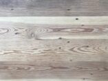 A wooden surface with visible grain patterns and natural variations in color. The wood displays a mix of light and dark tones, with some knots and lines running horizontally across the surface.