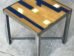 A small square table with a wooden top featuring a geometric design in blue and natural wood tones. The table has a metal frame and legs, and it is placed on a gray concrete floor.