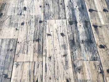A close-up view of a distressed wooden floor with a pattern of dark stains, scratches, and wear, highlighting the rustic and weathered texture of the planks.