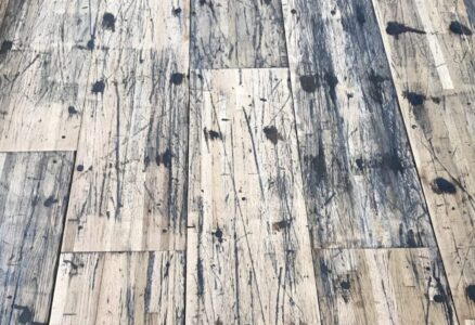 A close-up view of a distressed wooden floor with a pattern of dark stains, scratches, and wear, highlighting the rustic and weathered texture of the planks.