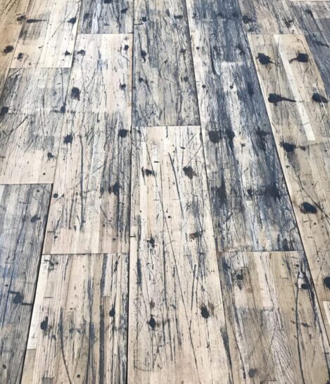 A close-up view of a distressed wooden floor with a pattern of dark stains, scratches, and wear, highlighting the rustic and weathered texture of the planks. A close-up view of a distressed wooden floor with a pattern of dark stains, scratches, and wear, highlighting the rustic and weathered texture of the planks.